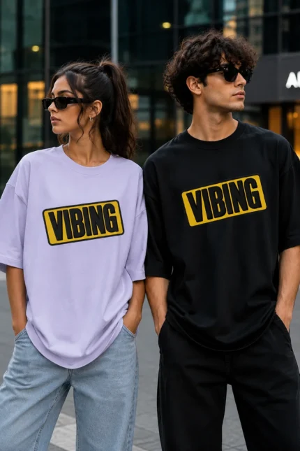 "Male and female models wearing oversized VIBING graphic T-shirts, featuring a lavender oversized tee for women and a black oversized tee for men, styled with casual streetwear in an urban city setting."